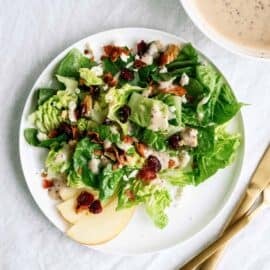 A plate of green salad with apple slices, cranberries, bacon bits, walnuts, and creamy dressing, beside a bowl of dressing and gold-colored cutlery on a white surface.