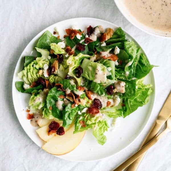 A plate of green salad with apple slices, cranberries, bacon bits, walnuts, and creamy dressing, beside a bowl of dressing and gold-colored cutlery on a white surface.