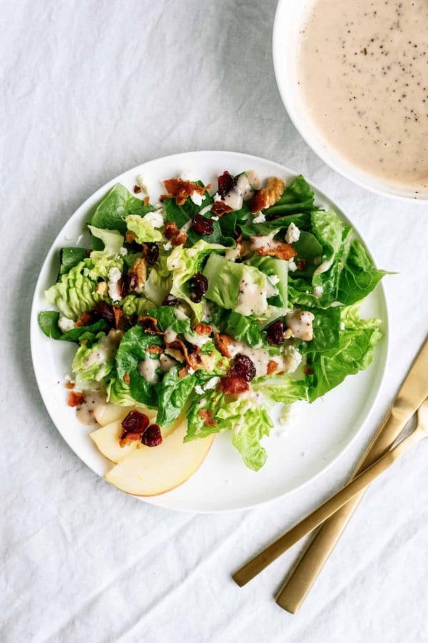 A plate of green salad with apple slices, cranberries, bacon bits, walnuts, and creamy dressing, beside a bowl of dressing and gold-colored cutlery on a white surface.