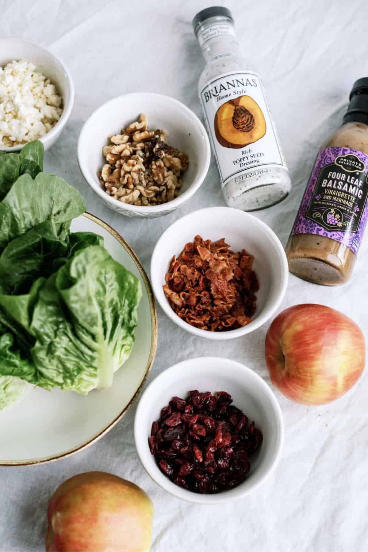 Assorted salad ingredients on a white surface, including lettuce, apples, walnuts, dried cranberries, crumbled cheese, bacon bits, and two bottles of salad dressing.