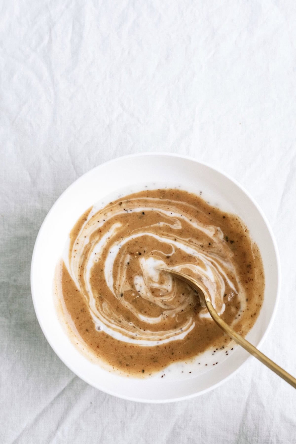 A white bowl with a spiral mixture of brown and white sauce, stirred with a gold spoon, on a white fabric background.