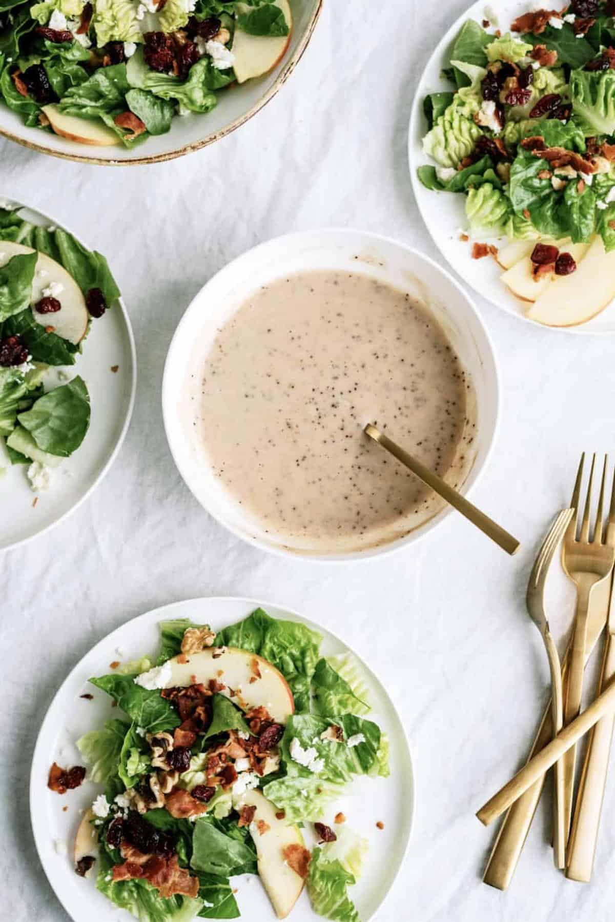 Four plates of salad with apple slices, lettuce, cheese, bacon bits, and dried cranberries surround a bowl of creamy salad dressing with a spoon; gold utensils are placed to the side.