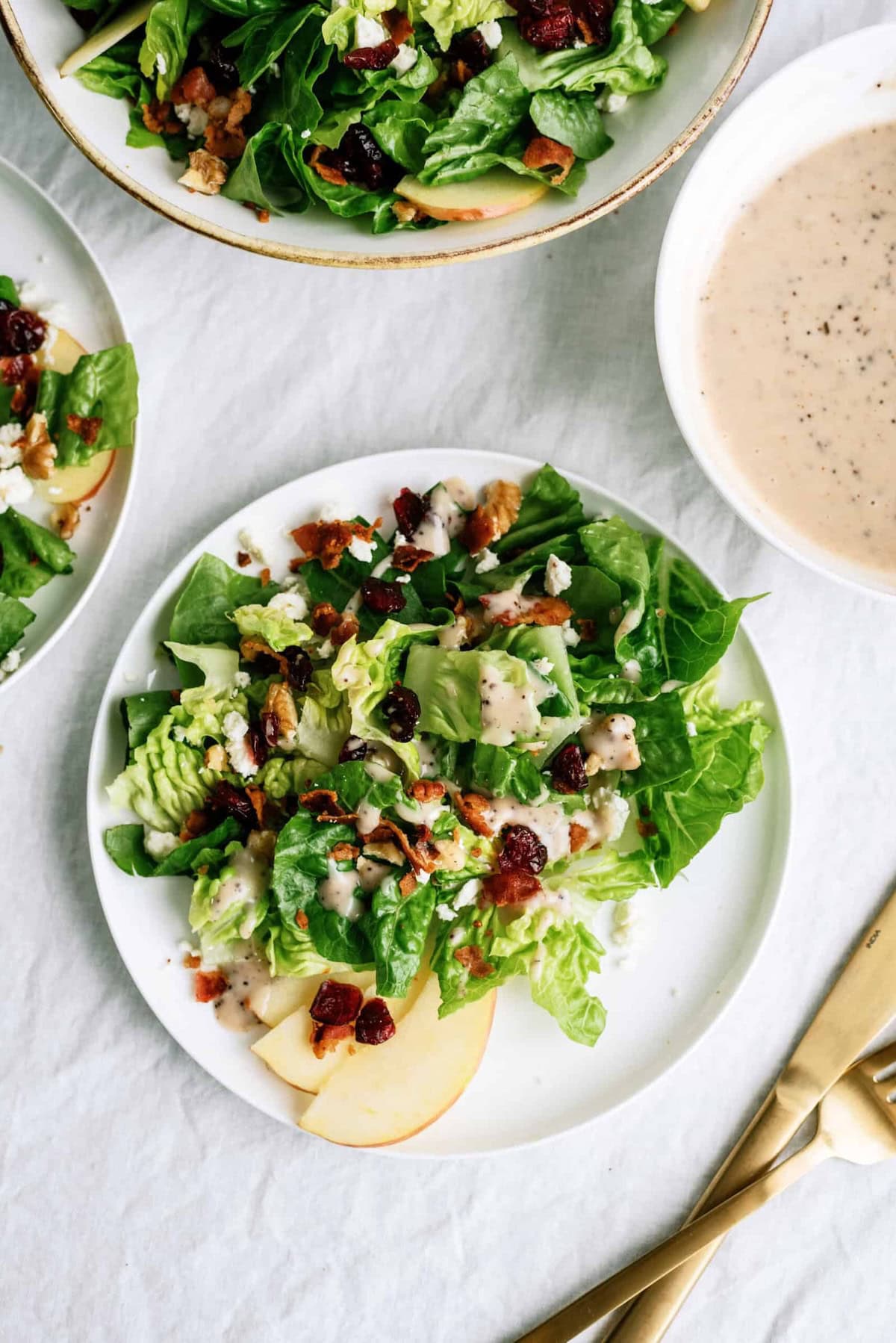 A plated salad with leafy greens, sliced apples, nuts, and dried cranberries next to a bowl of creamy dressing.