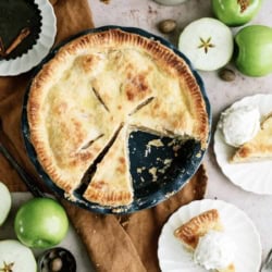 A baked apple pie with a slice removed sits on a table with green apples, spices, a brown cloth, and a plate holding a pie slice with vanilla ice cream.