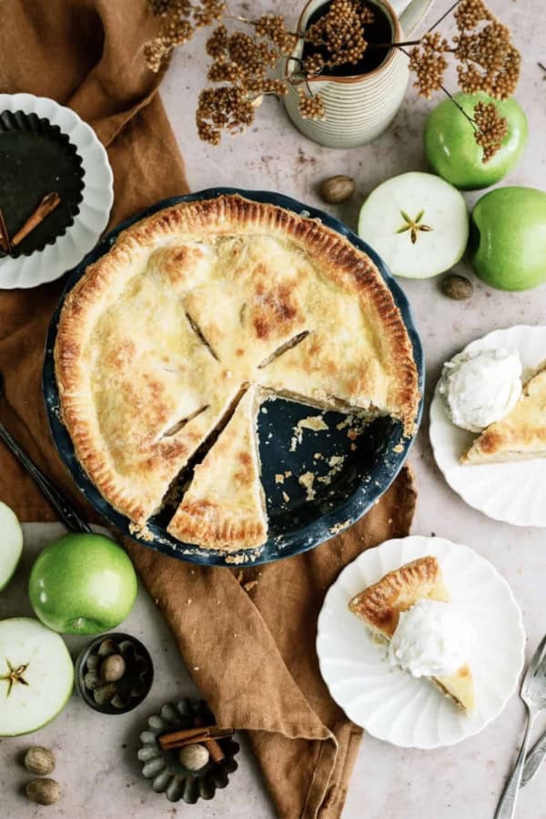 A baked apple pie with a slice removed sits on a table with green apples, spices, a brown cloth, and a plate holding a pie slice with vanilla ice cream.