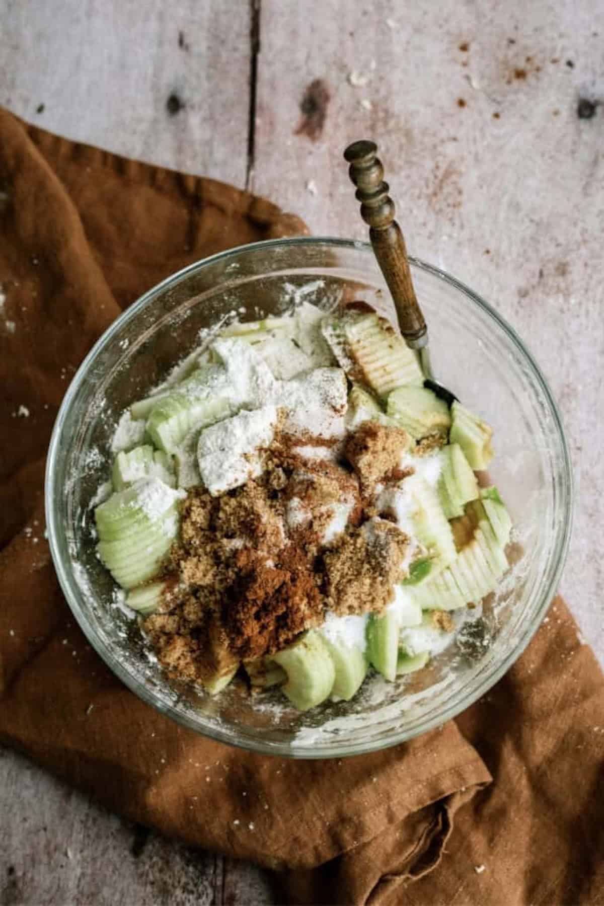 A glass bowl filled with sliced apples, flour, brown sugar, and cinnamon sits on a wooden surface with a brown cloth and a spoon.