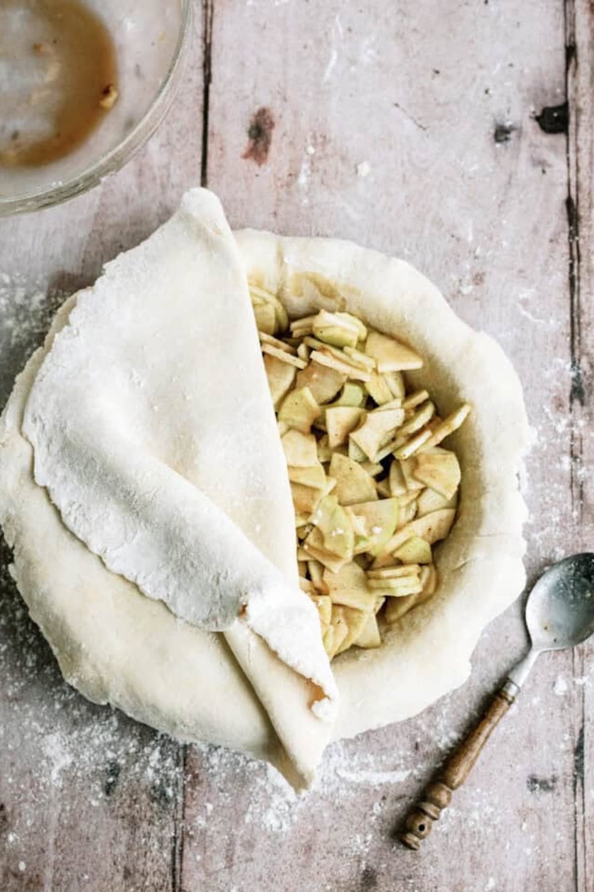 Unbaked apple pie with sliced apples in a crust, partially covered by a sheet of dough, on a floured surface next to a spoon and a bowl.