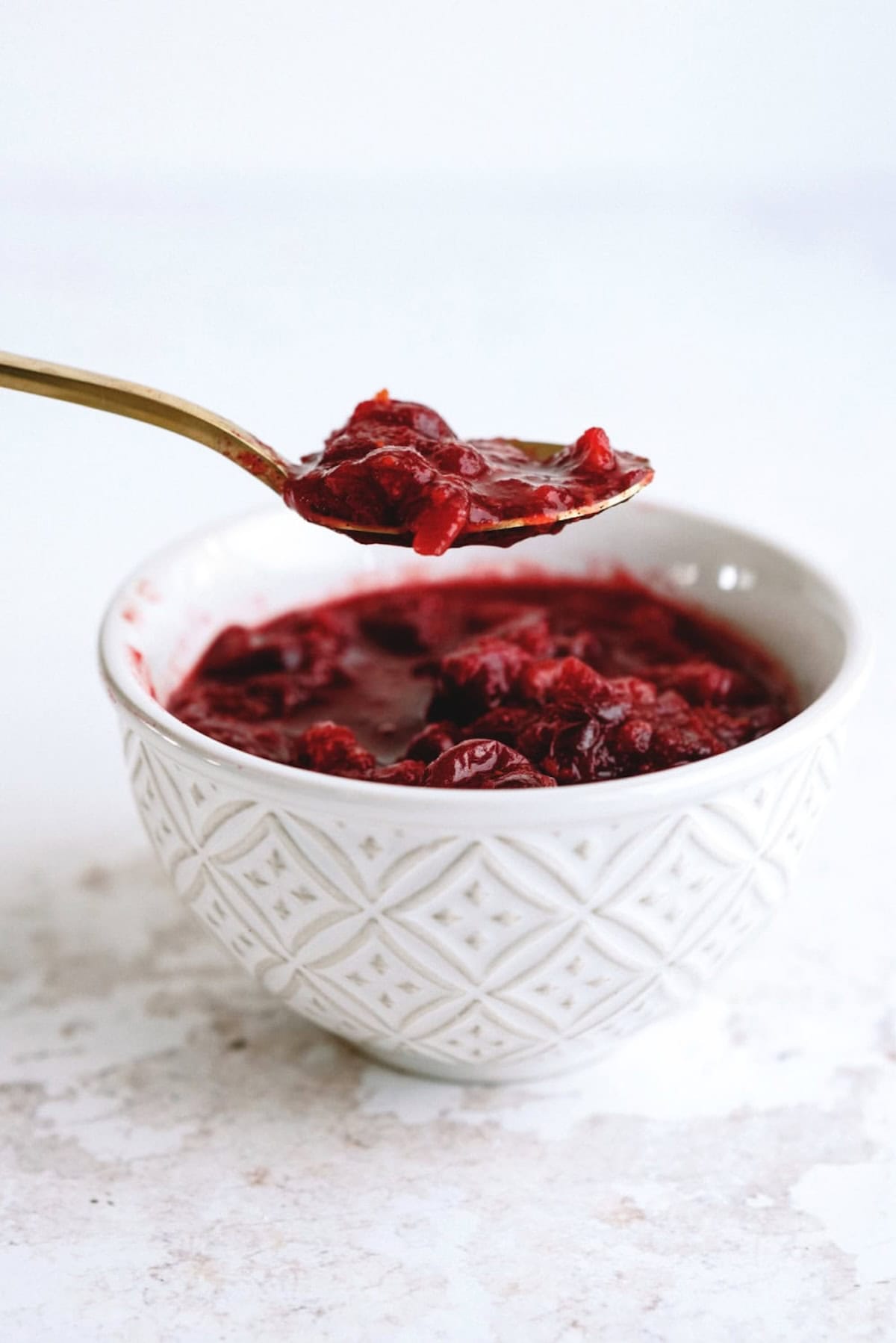 A spoonful of red cranberry sauce held above a white textured bowl filled with cranberry sauce.