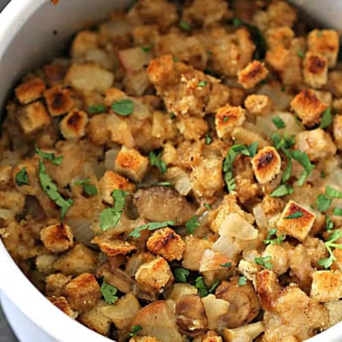 A white bowl filled with baked stuffing made of toasted bread cubes, onions, and herbs, garnished with chopped parsley.