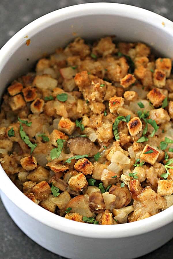 A white bowl filled with baked stuffing made of toasted bread cubes, onions, and herbs, garnished with chopped parsley.