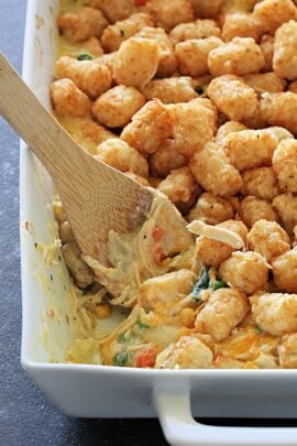 A close-up of a casserole dish filled with tater tots, vegetables, and creamy sauce, with a wooden spoon scooping a portion.