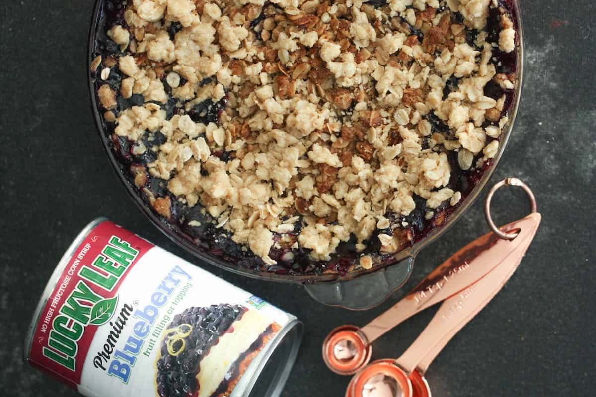 A baked blueberry crisp topped with oat crumble sits next to a can of Lucky Leaf blueberry fruit filling and measuring spoons on a dark surface.