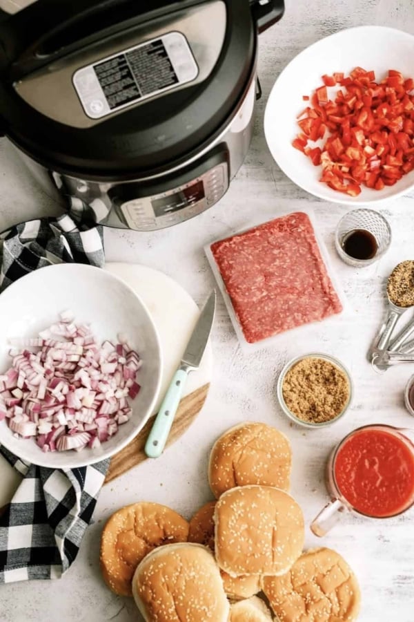 An Instant Pot, chopped red peppers, diced onions, ground beef, sauces, brown sugar, and burger buns are arranged on a countertop for meal preparation.