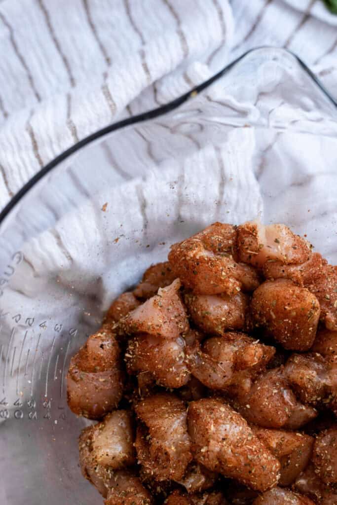 Raw cubed chicken in a glass bowl with seasoning.