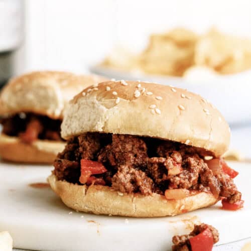 Sloppy Joe on a plate with another sloppy joe in the background and a bowl of chips.