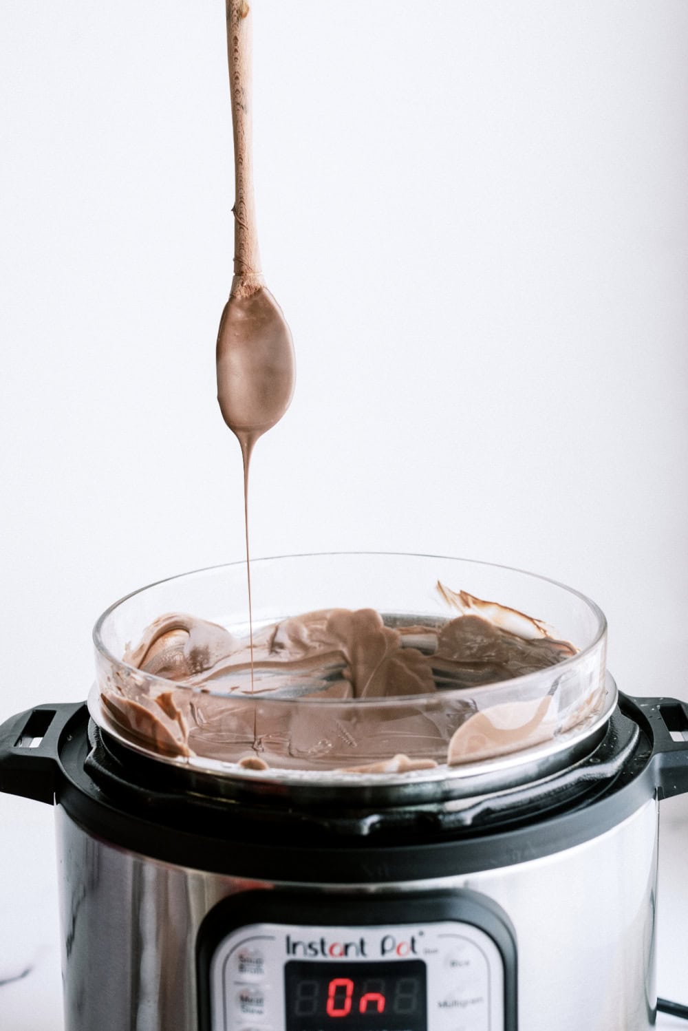 An instant pot with a glass bowl of melted chocolate. A wooden spoon held above the bowl dripping chocolate.