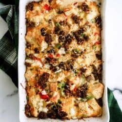 Baked casserole with bread cubes, bell peppers, onions, melted cheese, and browned sausage in a white rectangular dish on a green checkered cloth.