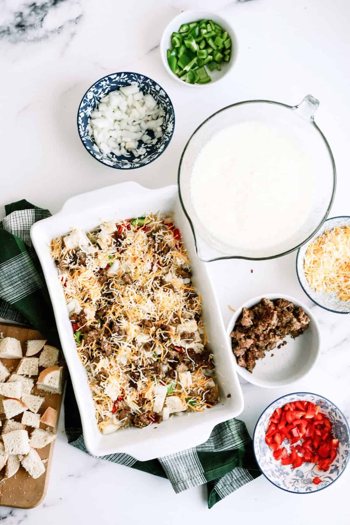 A casserole dish with ingredients like cheese, sausage, and peppers is surrounded by bowls of chopped onions, red peppers, green peppers, and bread cubes on a marble surface.