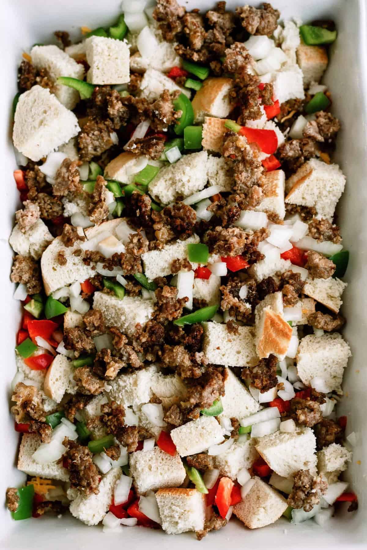 A baking dish filled with a mix of bread cubes, cooked ground sausage, diced red and green bell peppers, and chopped onions.