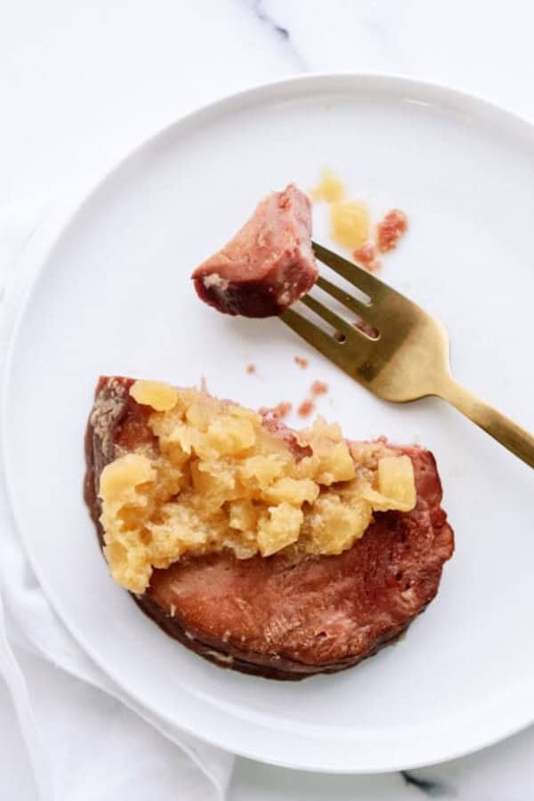 A slice of ham topped with cooked pineapple sits on a white plate next to a gold fork holding a small bite.