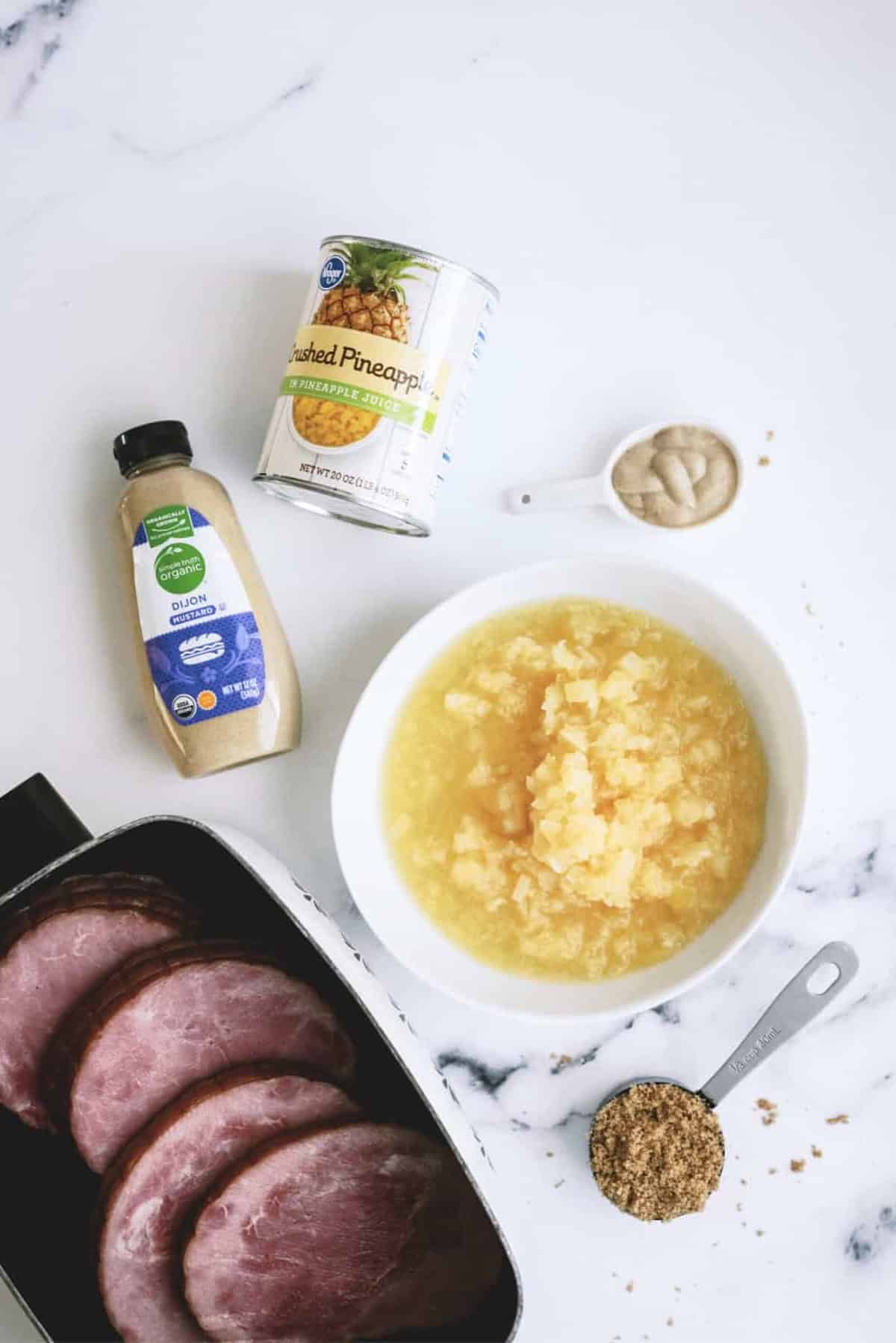 Sliced ham in a pan, a can of crushed pineapple, a bowl of pineapple, Dijon mustard, a spoonful of mustard, and a tablespoon of brown sugar on a white surface.