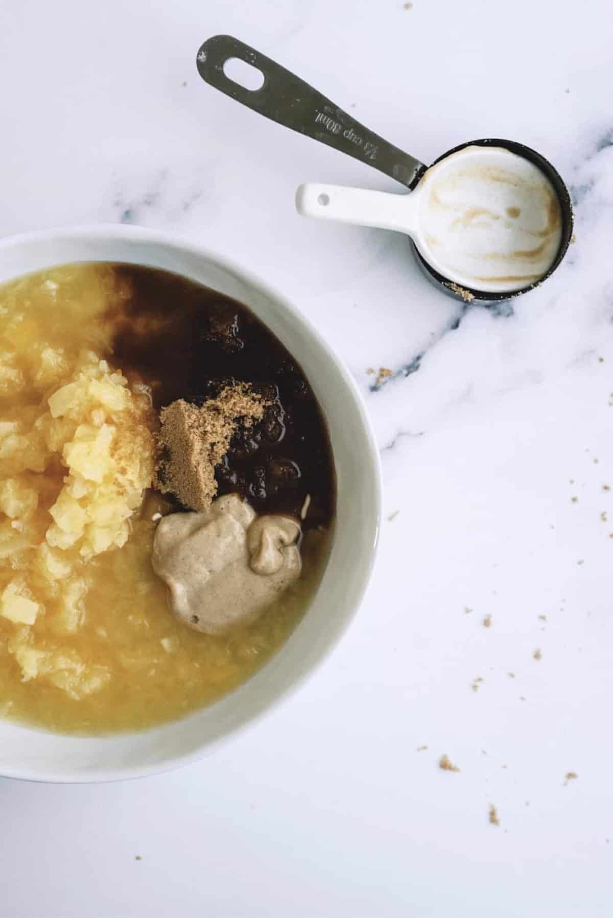A white bowl with crushed pineapple, brown sugar, spices, and a measuring cup with residue on a white marble surface.