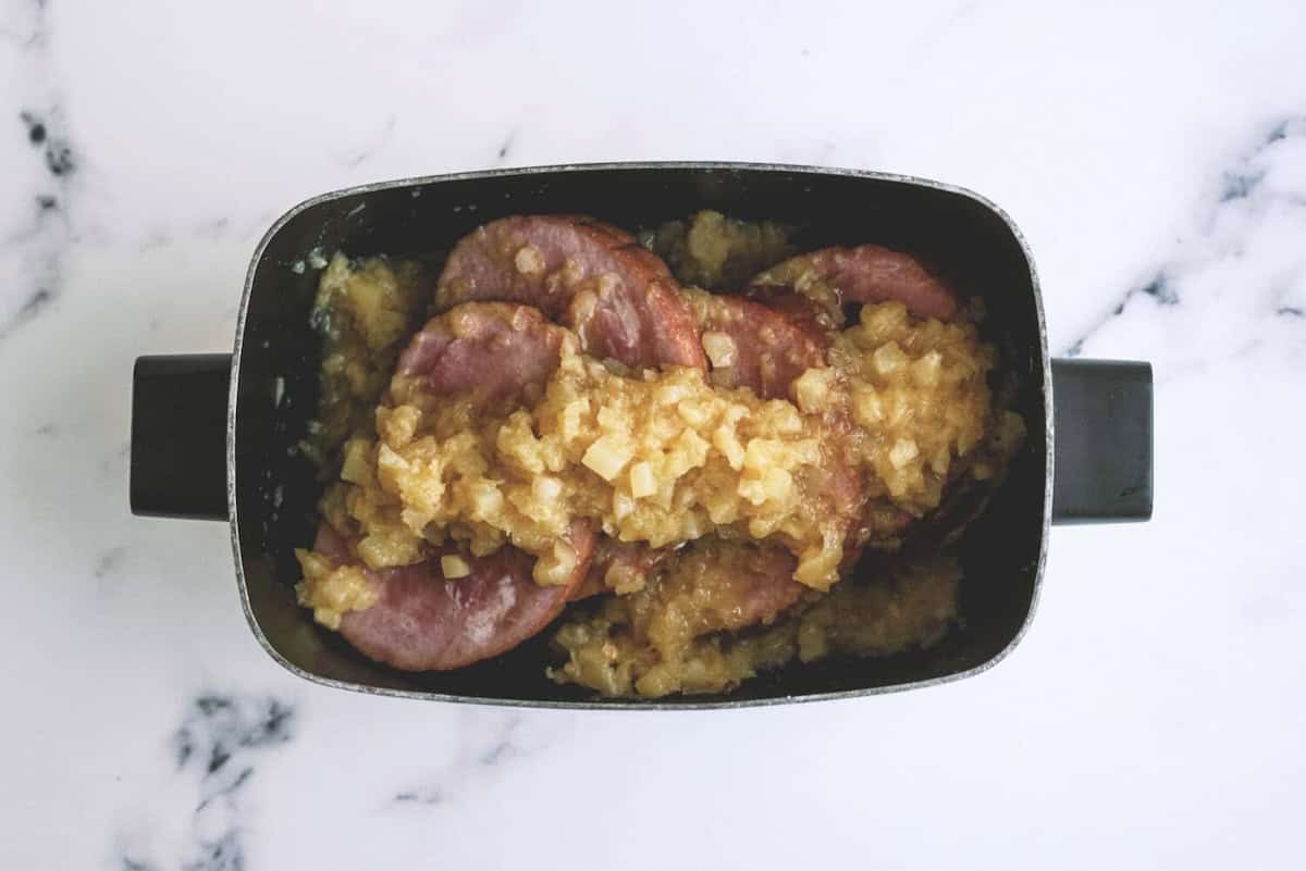 Sliced ham topped with a chunky pineapple and onion mixture in a black rectangular baking dish on a marble surface.