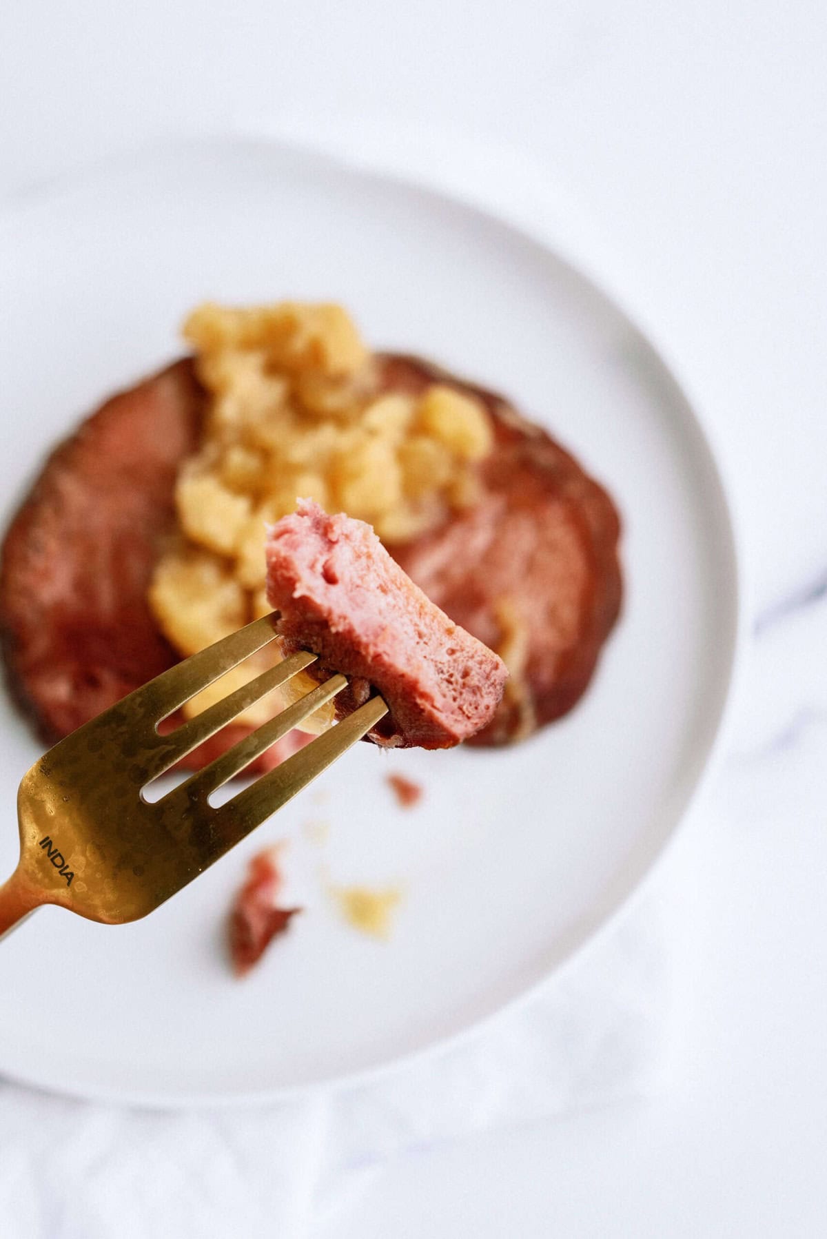 A close-up of a fork holding a piece of cooked ham, with a slice of ham and a pineapple topping on a white plate in the background.