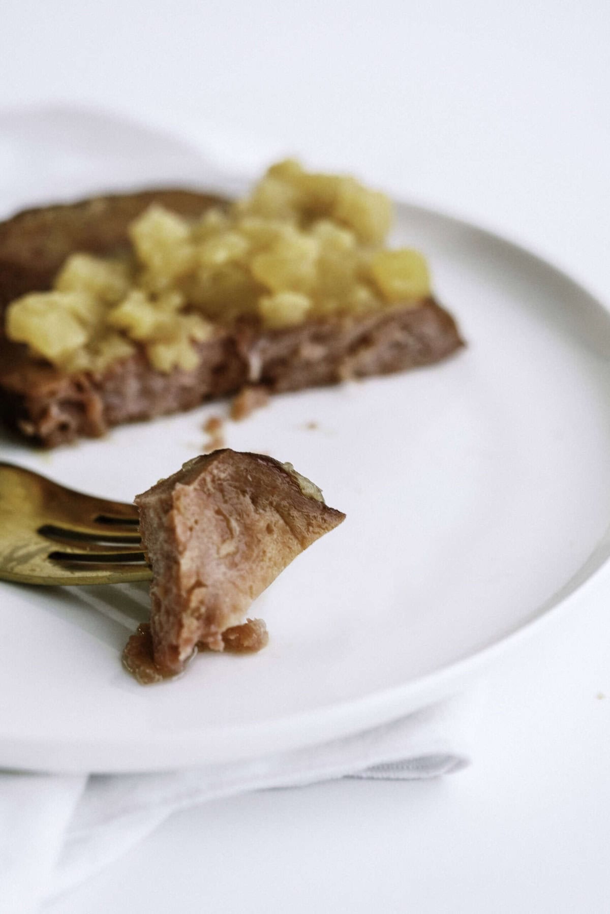 A close-up of a fork holding a bite of pork with pineapple topping on a white plate; the rest of the pancake is blurred in the background.