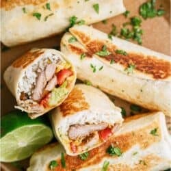 Four grilled burritos, two cut to show fillings of rice, grilled meat, guacamole, and vegetables, are served on a plate with chopped cilantro and a lime wedge.