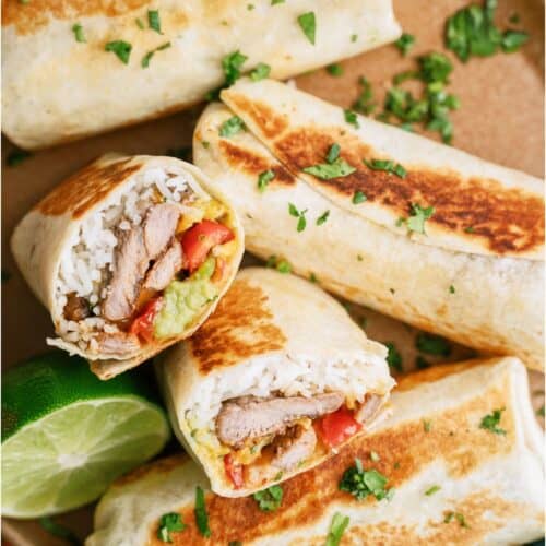 Four grilled burritos, two cut to show fillings of rice, grilled meat, guacamole, and vegetables, are served on a plate with chopped cilantro and a lime wedge.