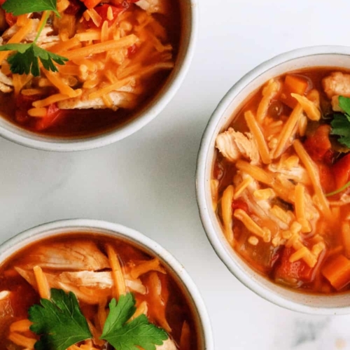 Three bowls of chicken chili topped with shredded cheddar cheese, diced vegetables, and parsley, arranged on a white surface.