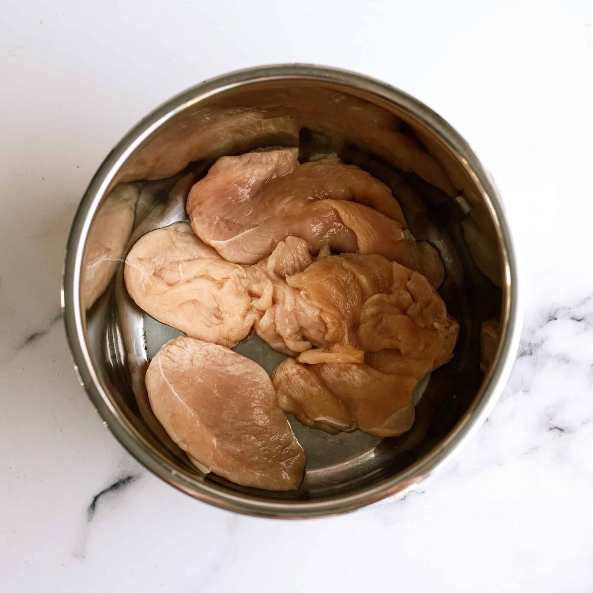 Raw chicken breast pieces placed in the bottom of a stainless steel pot on a white marble surface.