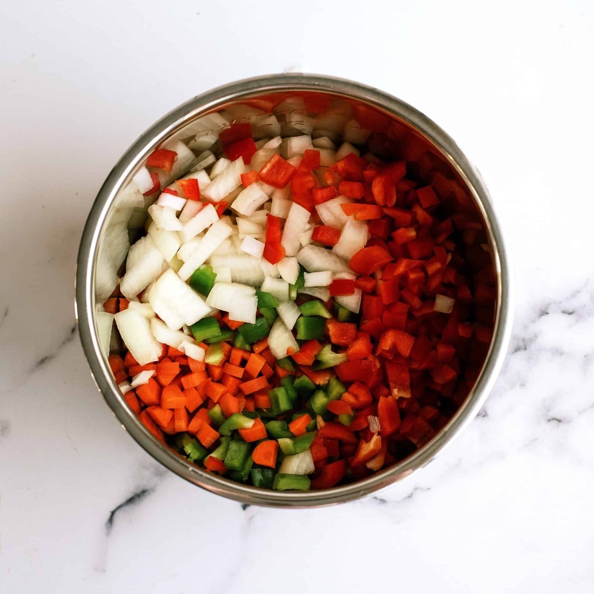 A stainless steel bowl filled with diced onions, red bell peppers, green bell peppers, and carrots on a white marble surface.