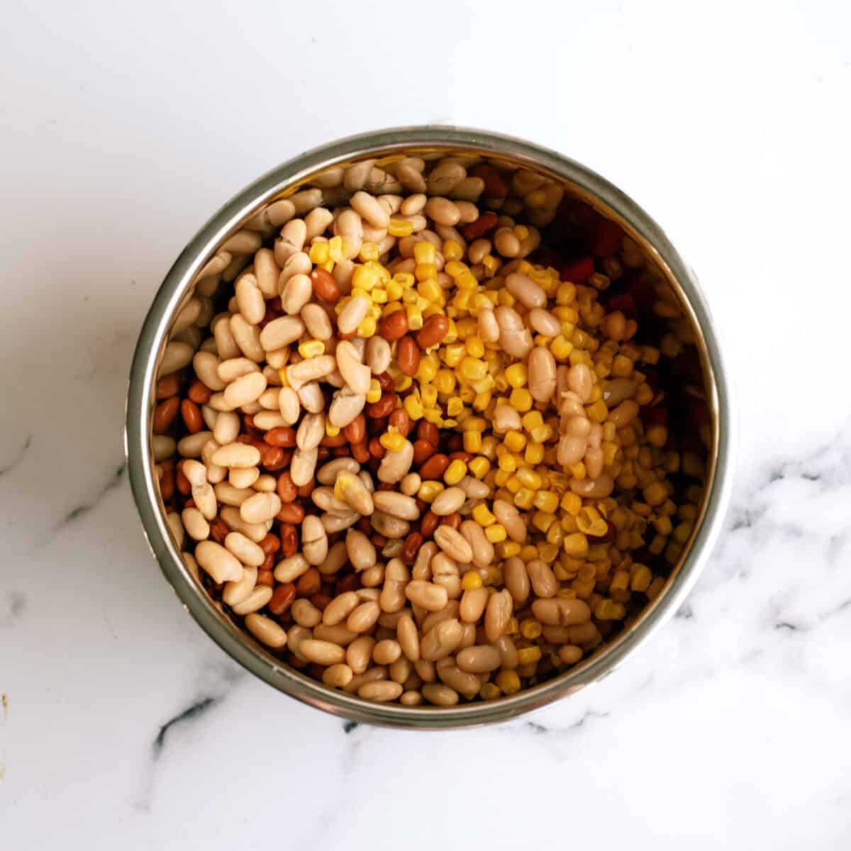 A metal bowl filled with white beans, kidney beans, and corn, placed on a white marble surface.