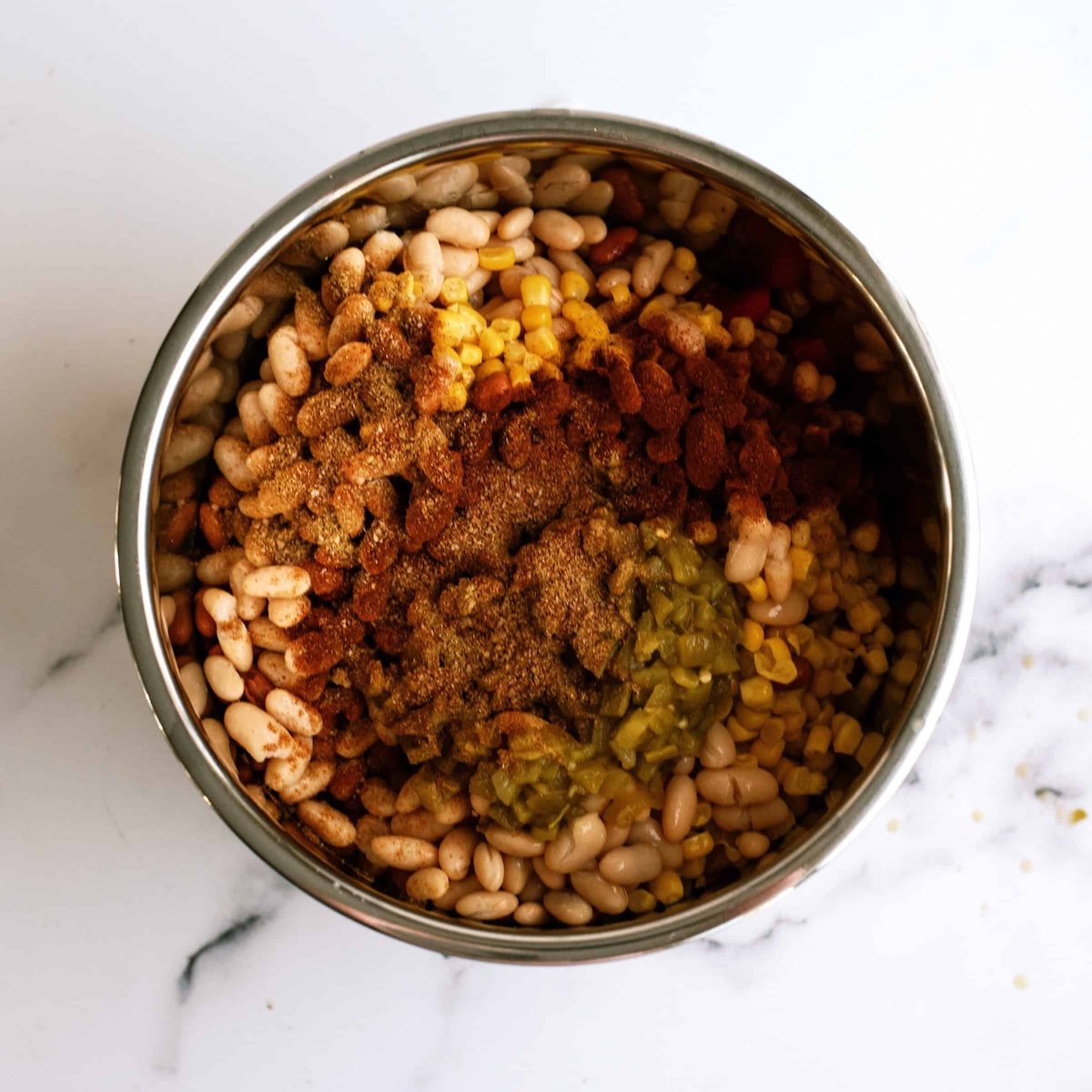 A stainless steel bowl containing white beans, corn, diced tomatoes, and seasoning on a white marble surface.