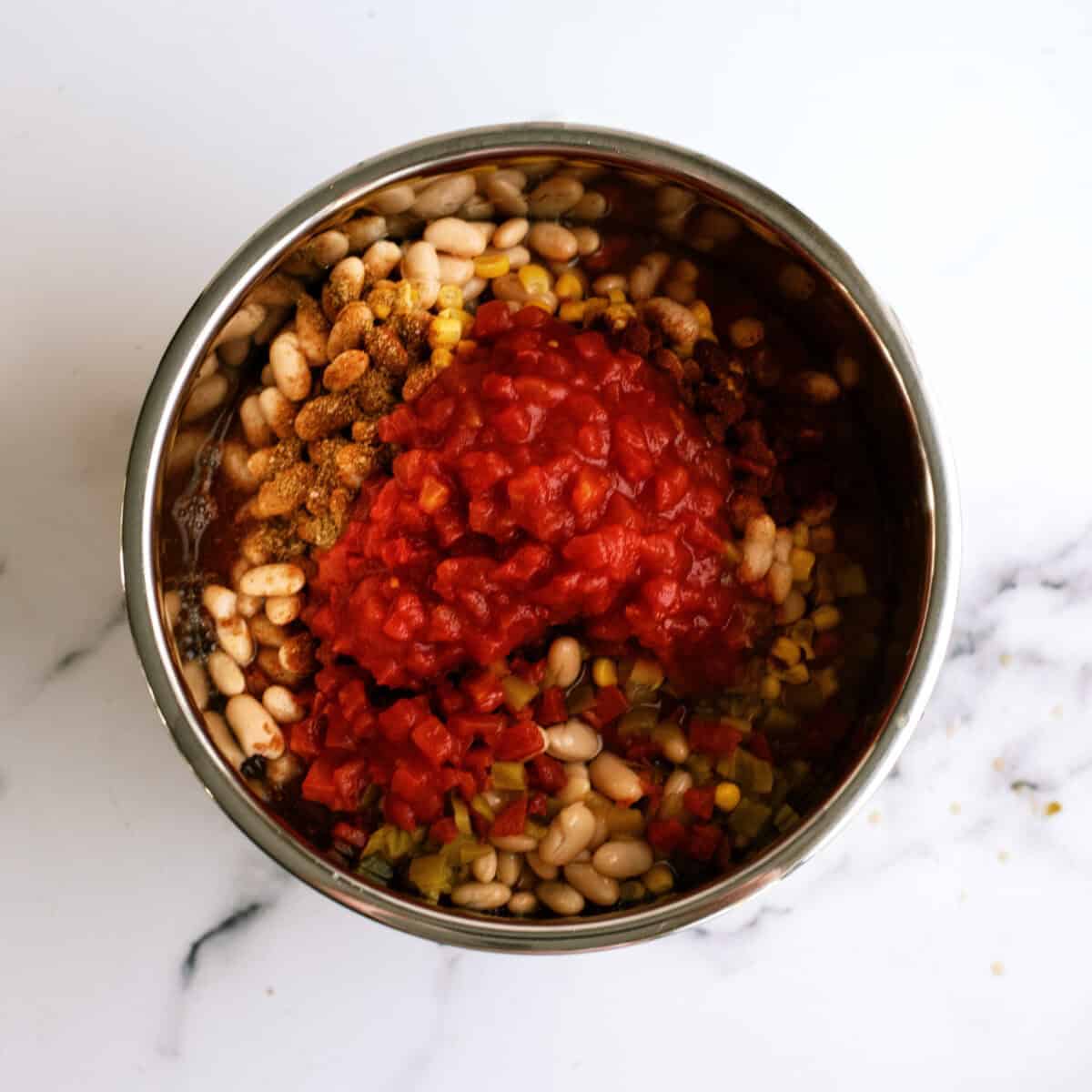A stainless steel bowl containing white beans, corn, diced tomatoes, and seasoning on a white marble surface.