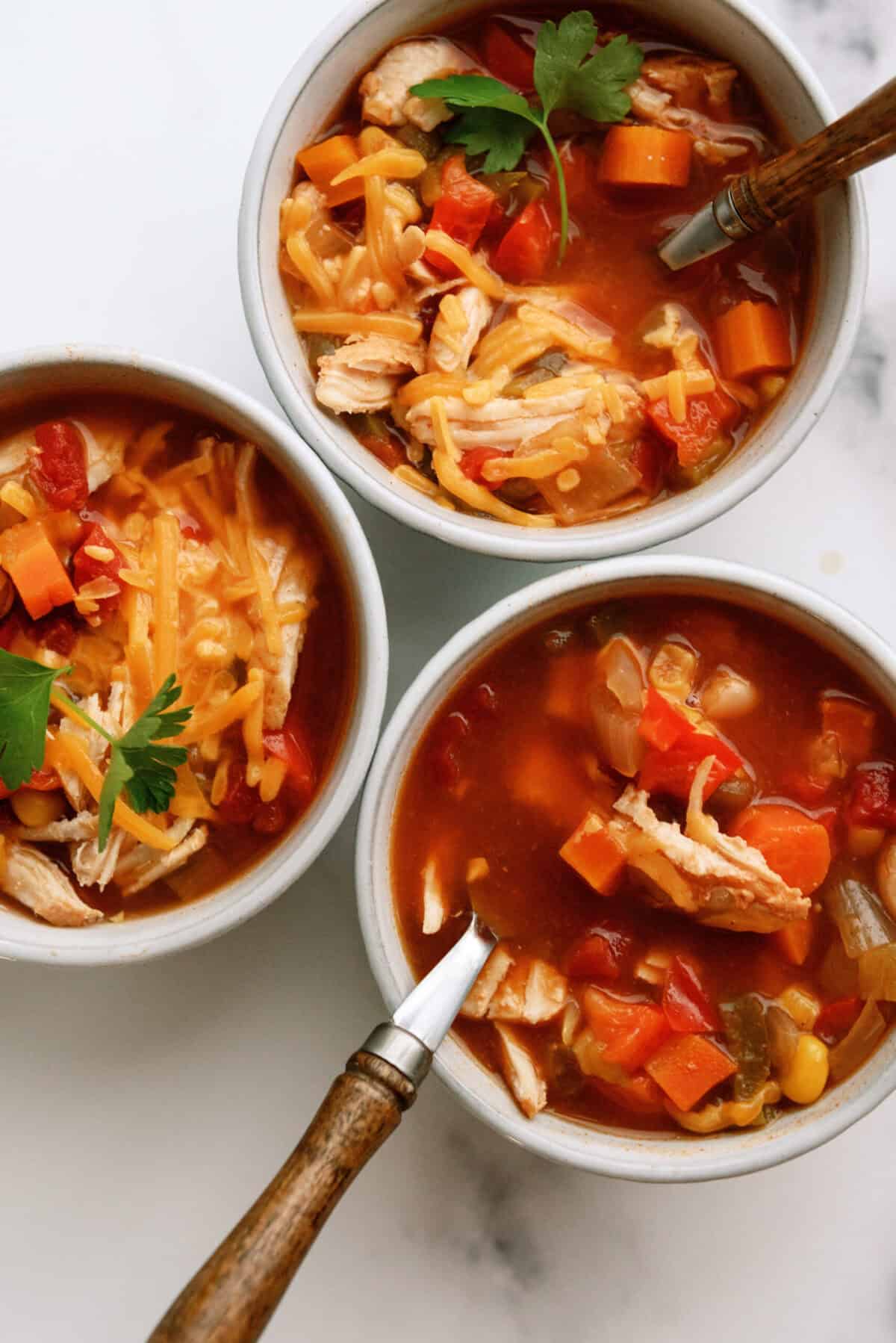 Three bowls of chicken chili garnished with shredded cheese and parsley, shown from above on a white surface with spoons in each bowl.