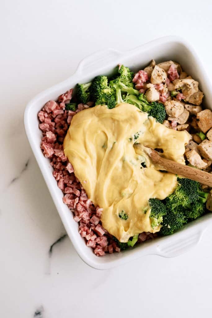 A square white dish contains chopped ham, broccoli, and diced chicken, topped with a generous layer of melted cheese sauce. A wooden spoon is partially visible in the mixture.