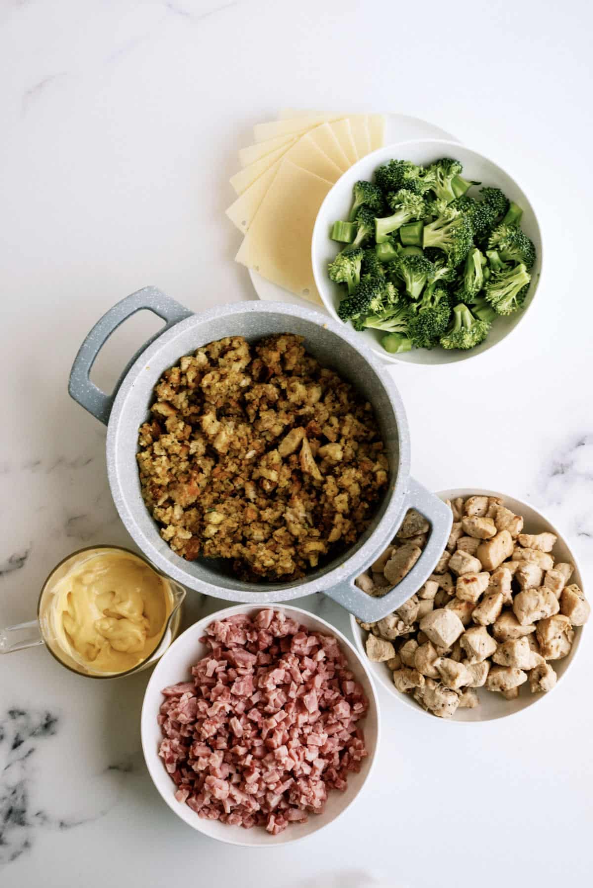 Ingredients for a casserole laid out on a table: chopped broccoli in a bowl, diced ham in a bowl, cubed chicken in a bowl, slices of cheese, melted cheese in a measuring cup, and stuffing in a pot.