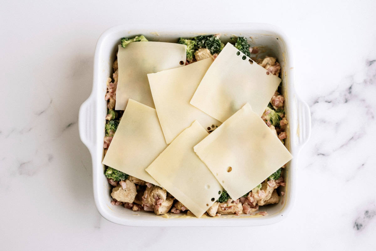 A square baking dish filled with a mix of chicken, broccoli, and cheese, topped with several slices of cheese, placed on a marble countertop.