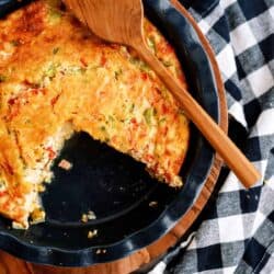 A partially eaten vegetable quiche in a round dish, placed on a wooden board with a wooden spatula on top, next to a black and white checkered cloth.