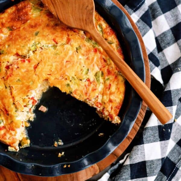 A partially eaten vegetable quiche in a round dish, placed on a wooden board with a wooden spatula on top, next to a black and white checkered cloth.