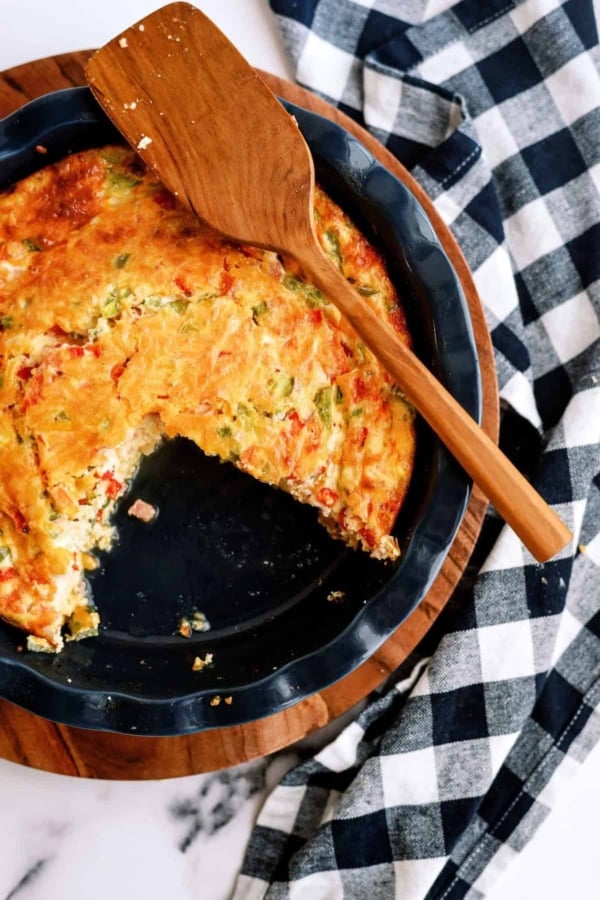 A partially eaten vegetable quiche in a round dish, placed on a wooden board with a wooden spatula on top, next to a black and white checkered cloth.
