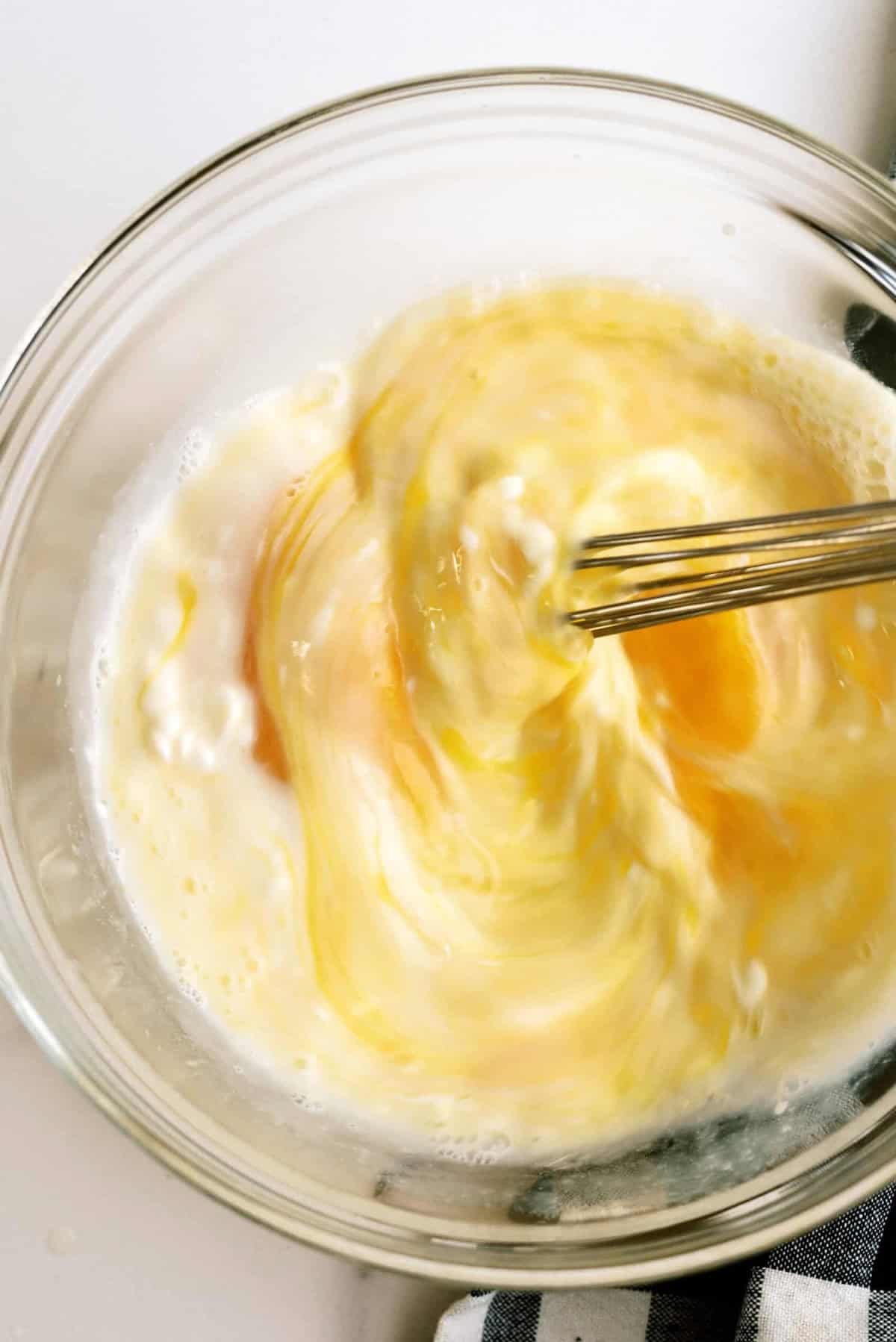 A whisk is beating eggs in a clear glass bowl, creating a mixture of yellow and white swirls.