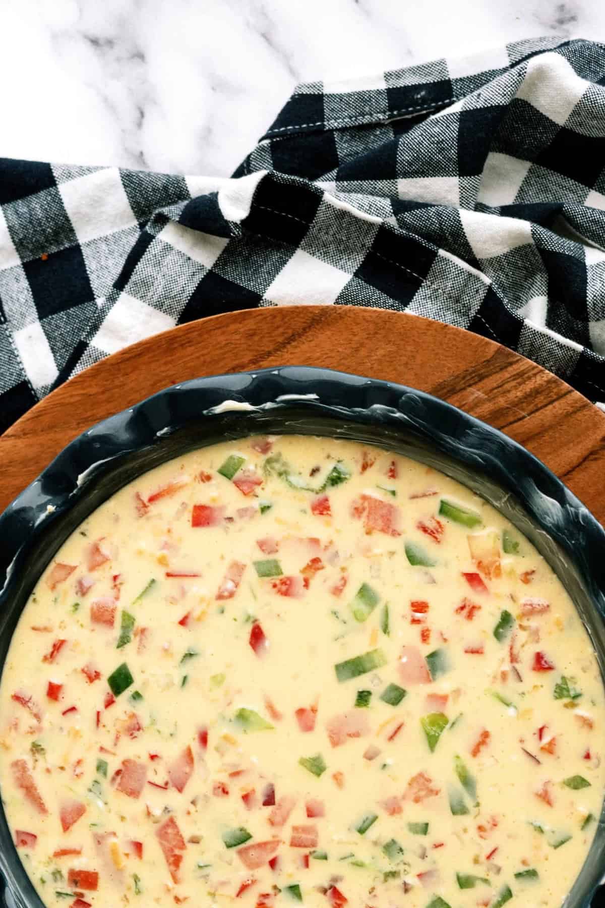 Unbaked quiche filled with diced red and green bell peppers in a black pie dish on a wooden surface, near a black and white checkered cloth.
