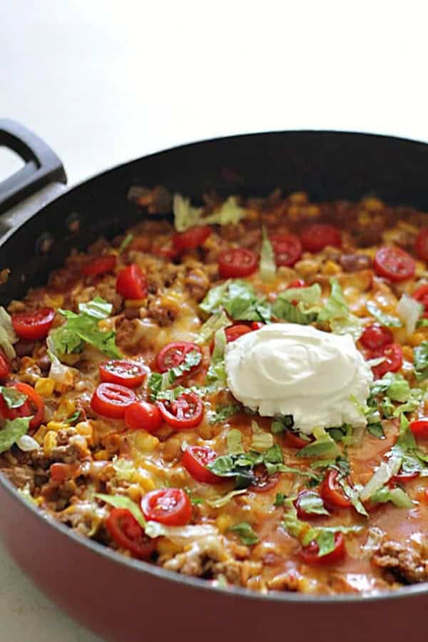 A skillet filled with a cheesy ground turkey dish topped with chopped lettuce, sliced cherry tomatoes, and a dollop of sour cream.