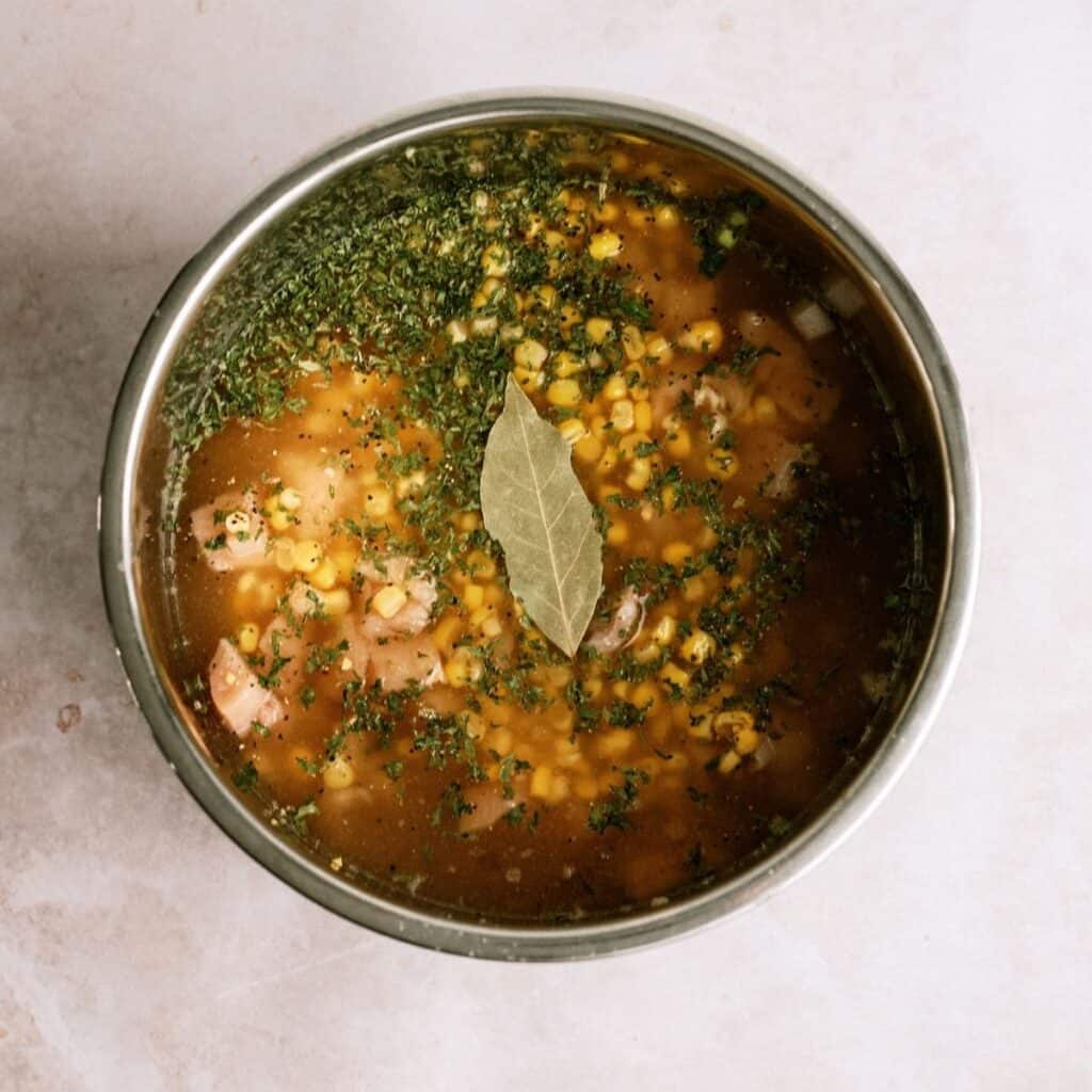 A pot of soup with corn, herbs, and a bay leaf floating on top, viewed from above on a light surface.