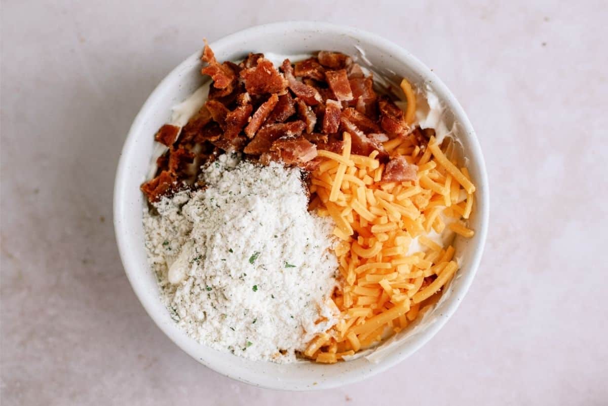 A white bowl containing chopped bacon, shredded cheddar cheese, and a white ranch mixture sits on a light-colored surface.