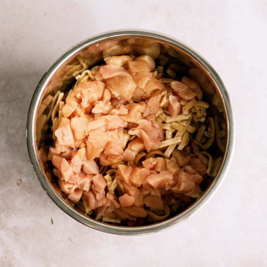 A stainless steel bowl filled with chopped raw chicken pieces and uncooked noodles on a light surface.